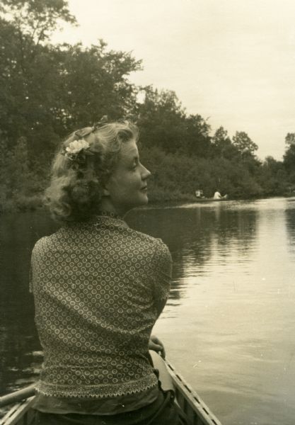 Mary Griggs (Burke), wearing a patterned short-sleeve blouse, shorts, and a flower in her hair, is sitting in the front of a canoe on the Namakagon River in Bayfield County. There is a canoe further ahead on the river.