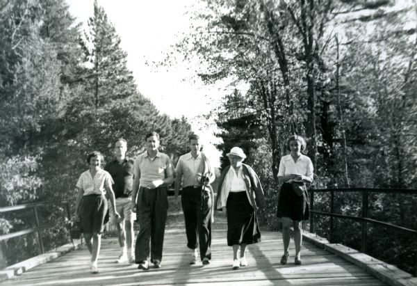 Mary Livingston Griggs and her daughter Mary are walking over the Brule River on a wood bridge with iron railings. Three men and one woman are walking with the Mary's and everyone except Mary Livingston Griggs is wearing casual clothes and short sleeves. Mary Livingston Griggs is wearing a long skirt, jacket and hat. Persons from left to right: Jane Matteson; Robert Matteson; Jackson Burke; Sumner Matteson; Mary Livingston Griggs; Mary Griggs Burke.