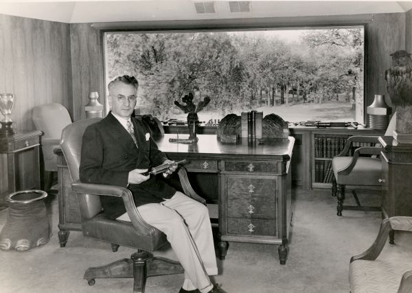 Oscar Zerk is sitting at his desk in his office holding a Zerk grease gun in his hand. There is a taxidermied owl on the cabinet on the right, and a large picture window behind the desk has a view of the grounds. Next to the desk on the left is an elephant foot cuspidor.