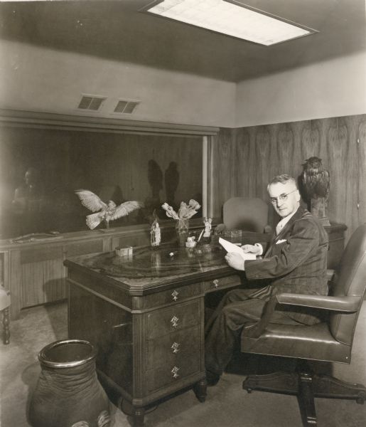 Oscar Zerk is sitting at a desk holding a paper in his hand. The room is decorated with two taxidermied birds, and a large picture window is behind the desk. Next to the desk on the left is an elephant foot cuspidor or wastebasket.