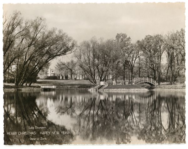 A holiday card featuring Lake Dunmovin on the Zerk estate. The mansion is in the background on the left. On the opposite shoreline on the right is an arched bridge leading to an island. On the island is outdoor furniture, steps to the water, and a diving board that extends into the lake. Text at bottom left reads: "Lake Dunmovin, Merry Christmas Happy New Year, Oscar U. Zerk".