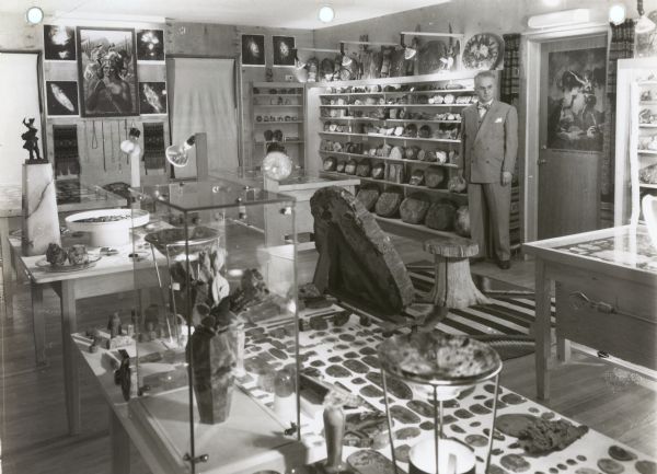 An exhibit of petrified wood sections and other objects on tables and display cases in the Zerk Museum. Paintings, photographs, jewelry and other objects are displayed on the walls and shelves surrounding the room. Oscar Zerk is standing by the door.