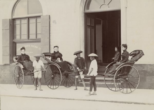 Mrs. Waldo, Mrs. Eliza Scidmore (in center holding a puppy in her lap), and an unidentified woman each sitting in rickshaws, which are also known as jinrikisha. Three Asian men are assisting them. Behind them a woman is standing in the doorway of a building which has a sign in the window that reads: "Canadian Pacific Railway C<u>o.</u>". Caption reads: "Mrs. Waldo. Mrs. Scidmore. M. O. No. 14 — Bund - June 1898".