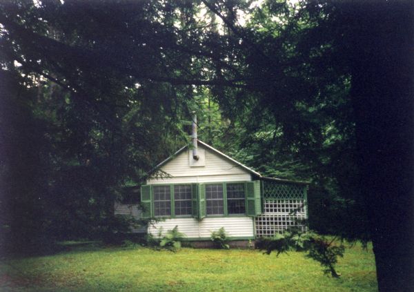 A white playhouse with green trim, windows and shutters situated in the forest. A sliding wood lattice gate covers the front porch, and a stove pipe extends above the roof.
