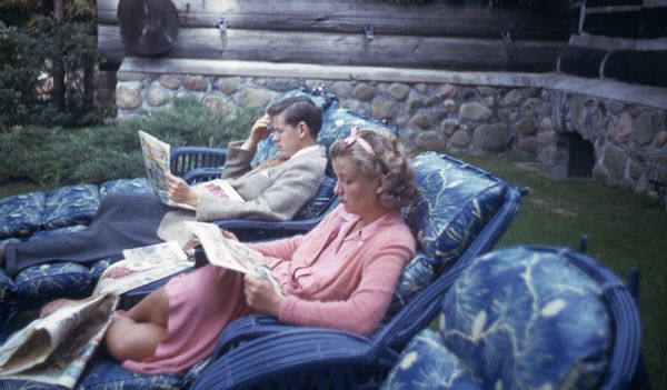 Mary Griggs, wearing a pink dress, cardigan and hair bow, and a male friend are lounging on blue painted wicker chaise lounge chairs with blue floral cushions in the back yard of the Main Lodge; both are reading the Sunday newspaper. The young man is dressed in blue slacks a gray jacket, and blue argyle socks.