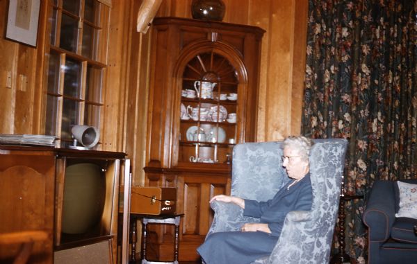 Mary Livingston Griggs, wearing a blue skirt and jacket, is seated in a pale blue brocade wing-backed chair, facing a television in the great room of the guest house. A built-in corner china cabinet with porcelain dishes is next to Mary and a window floral print drapes is behind Mary.  A phonograph is on a spindle-legged table next to the television. The arm of a dark blue with red piping upholstered sofa can be seen behind Mary. The entire room is wood paneled.
