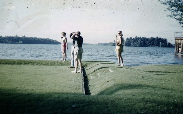 Two couples playing English lawn bowling on Forest Lodge bowling green next to Lake Namakagon. One woman is wearing pink Bermuda shorts and a white sleeveless blouse, the other woman a short, yellow-checked summer dress. Champagne Island is in the background as well as a small portion of the boathouse.