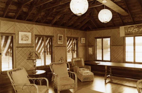 Interior of the second floor of the Forest Lodge boathouse. Bamboo-type wall paneling hung with numerous bird prints. Rattan chairs with cushions and a long wood table. Ball-type ceiling lamps hang from the ceiling; a table lamp and a lampshade with birds sits on a side table.