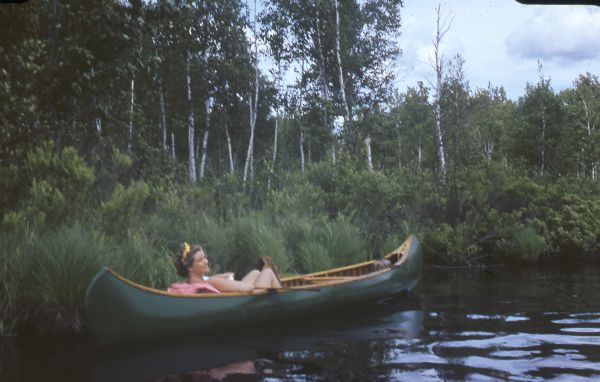 Mary Griggs is lounging in a cedar strip canoe with green canvas covering, along the shoreline of the Namakagon River. Mary has a yellow bow in her hair.