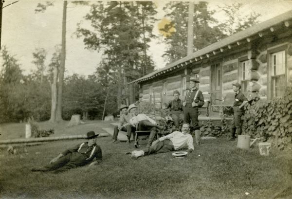 Crawford Livingston, wearing white shirt with a straw boater hat laying on the ground in front of him, and six friends are in various longing positions in front of a vertical log one-story structure at a former logging camp. A few large pine trees and some stumps are around the lawn. Some foliage, possibly hollyhocks, are growing along the base of the log cabin. In 1902, the log cabin became the main lodge at Forest Lodge when Crawford Livingston purchased the property.