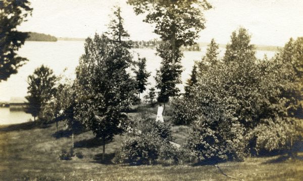 Woman wearing a white dress is standing among trees on a hillside that slopes towards Lake Namakagon. Champagne Island is seen in the background.