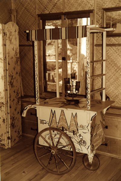 Three wheel wood cart with a handle and four upright wood struts that form an open roof.  The sides are painted with an alpine-type scene. A candelabra sits in the center of a painted star on the cart top. The cart is sitting in front of a window on the second floor of the boathouse.