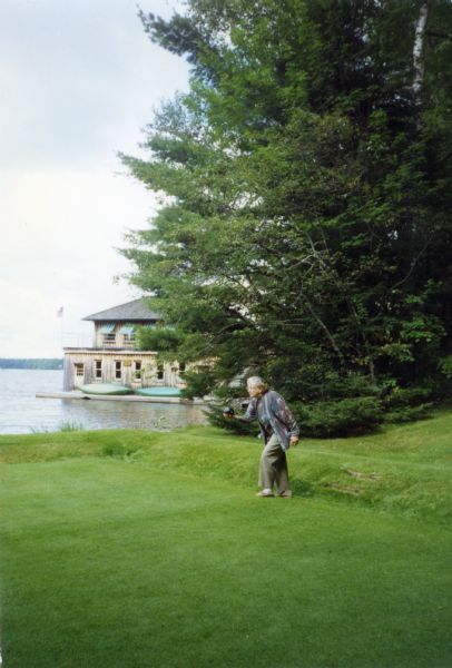 Mary Burke is standing on the  grass bowling green, preparing to toss a black ball down the pitch during the 100th anniversary celebration for Forest Lodge. She is wearing gray slacks and a printed cardigan sweater combination. Large evergreen trees are in the background. The boathouse with two green canoes on the deck can be seen beyond the trees.