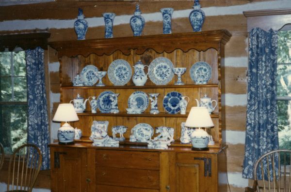 Blue and white porcelain dishes, vases, figurines and lamps on a wooden side-board set against a chinked log wall. Windows with blue and white curtains topped with wood valance's are located on each side of the side-board. Two spindle-backed chairs are in front of the windows.