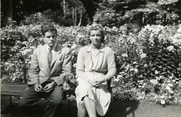 Mary Griggs at Forest Lodge, sitting on wooden bench in front of flower garden with her pet dachshund and a male friend. Mary is wearing a dress with a cardigan, the male friend is wearing a sport jacket.