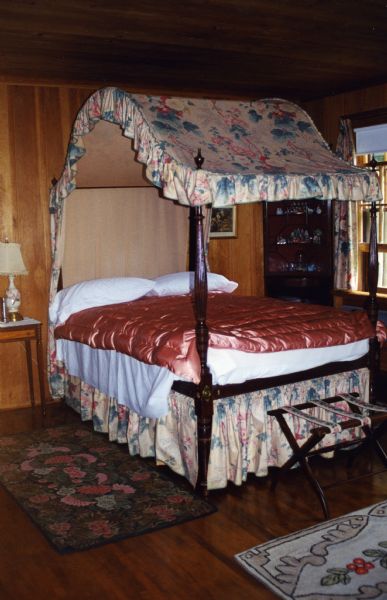 Pink ruffled canopied wooden bed with salmon pink quilt and a suitcase stand at the foot of the bed. There is a corner cabinet with glassware and a bedside table with a porcelain lamp. Two hooked rugs are on the wood floor, and there's a partially open window.