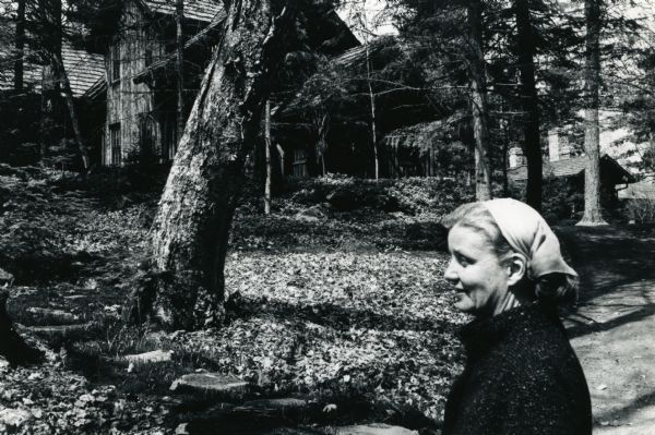 Mary Griggs is outdoors wearing a kerchief and coat. Forest Lodge and the grounds are in the background.
