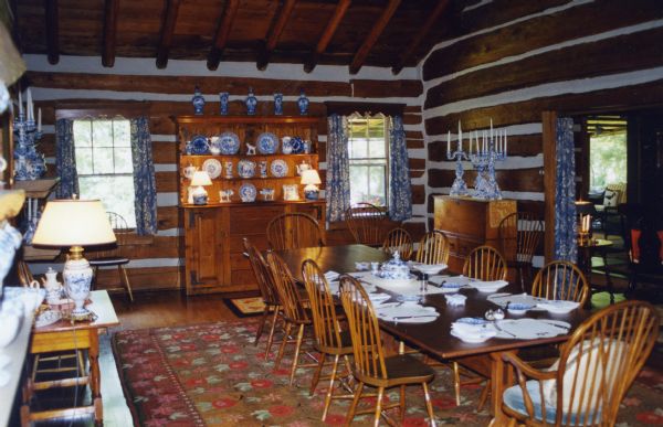 The dining room in the main lodge, with chinked log walls, wood ceiling beams, two curtained windows and a curtained doorway leading to the living room. In the center of the room is a wood dining table with Windsor chairs situated on a large area wool rug. The table is set for seven with Royal Copenhagen china. Three Royal Copenhagen candelabra's are on top of a wood storage cabinet alongside one log wall. Around the remaining log walls are assorted Royal Copenhagen dishes, creamers and lamps.