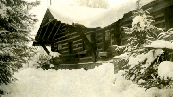 Horizontal chinked log building with a stone foundation and wood window boxes. The roof, grounds and surrounding spruce trees are covered in snow.