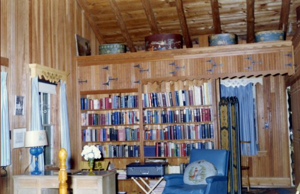 Wood-paneled room with exposed log ceiling joists. One wall of the room has an all wood bookcase full of books, and above are upper doors to cabinets with four hatboxes on top. There is a blue leather easy chair with a pillow, and a small blue suitcase on a stand in front of the bookcase. A desk with a vase of white flowers and a blue lamp is between a window and a door that leads outdoors. Under the shelf with hatboxes is a folded privacy screen.