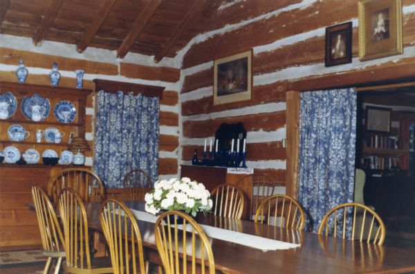 A wood dining room table with a flower bouquet is surrounded by wooden Windsor chairs in a chinked log room with a log beam ceiling. Royal Copenhagen blue dishes are on a wood hutch, and blue glass candlesticks are on top of another wood sideboard. Blue and white curtains cover a dining room window and a doorway leading into the living room. Framed images hang over the doorway.