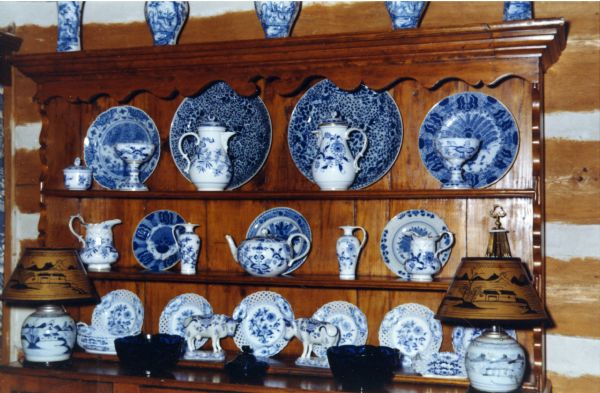 An assortment of Royal Copenhagen blue dishes are arranged on the upper open shelves of an antique wood buffet in the dining room of the main lodge. The collection includes plates, creamers, pitchers and urns, vases, teapots, and lamps. There are also three midnight blue glass bowls sitting on the buffet between the lamps.
