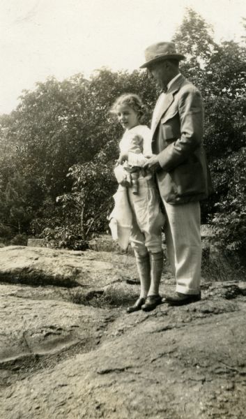 Theodore Wright Griggs and his daughter Mary are standing on rocky ground with woods in the background. Mary is wearing a shirt, knickers, knee-socks and shoes while holding a doll. Theodore Griggs is wearing a sport jacket, bow-tie, cuffed trousers, and shoes.