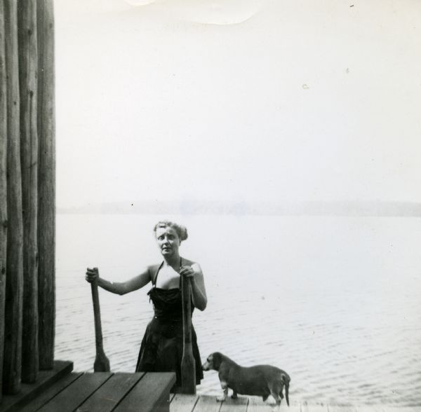 Mary Griggs (Burke) is wearing a one-piece bathing suit as she climbs out of Lake Namakagon onto the Forest Lodge boathouse deck while her dachshund awaits.