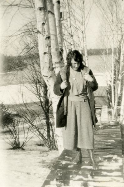 Mary Livingston Griggs is standing on the birch-lined boardwalk leading to the Forest Lodge boathouse. Mary is wearing a wool dress, coat and leather boots. Lake Namakagon is in the background.