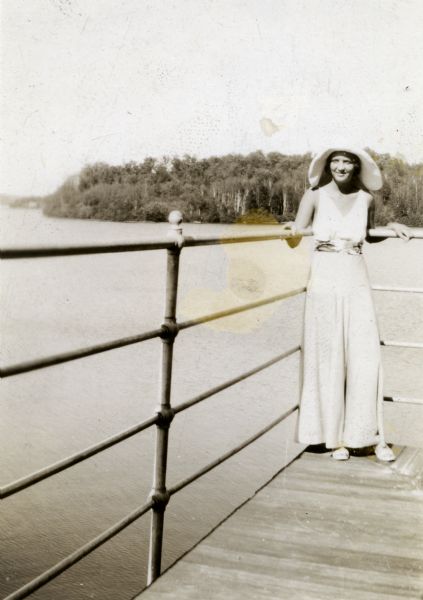 Mary Griggs (Burke) is leaning against a pipe railing on the corner of the second story of the Forest Lodge boathouse. Mary is wearing a wide-legged jumpsuit and a floppy hat. Champagne Island is in the background.