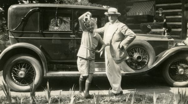 Mary Griggs and her father Theodore W. Griggs are standing next to a four-door automobile parked outside the main lodge at Forest Lodge. Mary's mother, Mary Griggs, is standing in the background on the front porch. Young Mary is wearing shorts, blouse, shoes and socks while holding a cap over her head. Her father is wearing a summer suit and hat, and they both have their left hand on their hips. The automobile has a hood ornament and spoke-wheel tires with a spare tire stored on the running board behind the front fender. Iris flowers are growing in the foreground. The photographer is reflected in the window of the car.