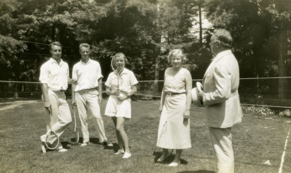 On Forest Lodge badminton court, left to right: Al Luideke, George Meine, Mary Griggs, Harriet Thompson, and Mary's father, Theodore Griggs who is addressing the four badminton players. Theodore is wearing a white suit and is holding his hat. The two young men wear white trousers and short sleeve shirts, Al leans on his racket as his right ankle is crossed over his right; Mary is wearing baggy white shorts and a white short sleeve blouse as she hold her badminton racket if front of her face; Harriet is wearing a long white skirt and a sleeveless top. Evergreen trees grow in the background. 
