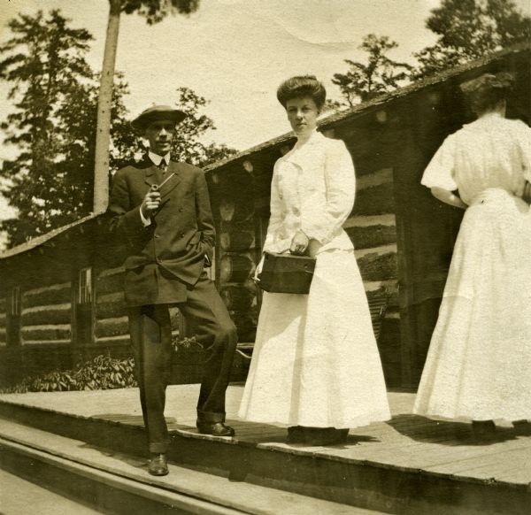 Mary Livingston and W. Blair Flandreau are standing on the porch of the original Forest Lodge, along with another woman with her back to the camera. Mary is dressed in a long two-piece white dress with her hair in an up-do as she holds a camera, possibly a No. 4 Cartridge Kodak. Blair is wearing a double-breasted suit and a hat as he stands with one foot on the porch and the other on a lower step as he smokes a pipe.