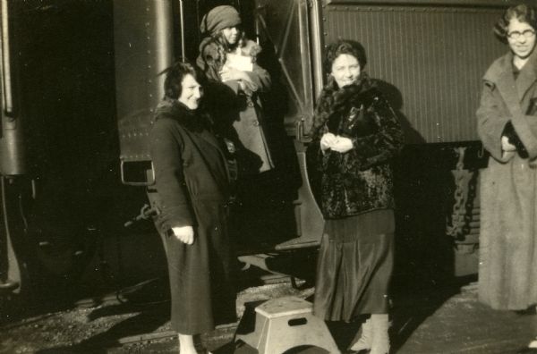 Mary Livingston Griggs, her daughter Mary, and two unidentified women are preparing to board a train car. Young Mary is in the open train  door, wearing a buttoned winter coat and hat as she holds her doll. Mother Mary is standing on the ground next to the train door, wearing a fur coat over a three-quarter length skirt, and wearing spats. An unidentified woman who is possibly pregnant and likely a relative, is standing on the other side of the open train door wearing a wool coat with a fur collar. An unidentified younger woman, probably young Mary's nurse, is wearing a wool coat and has eyeglasses.