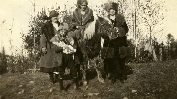 Mary Livingston Burke is sitting on Mary's pony as daughter Mary and two unidentified women are standing alongside. Daughter Mary is wearing a wool coat with a fur collar, wool knickers, knee-socks and shoes, a wool cap with a feather, and is holding a white cat. Mother Mary is wearing a wool coat with a fur collar and leather gloves. The other two women are wearing wool coats with fur collars and wool hats. The brown pony has a white mane.