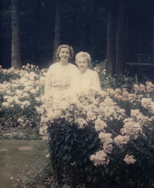 Mary Griggs and her daughter Mary are standing outside of the Forest Lodge guesthouse in a phlox flower garden.  Both Marys are wearing white dresses. A corner of the guest house screen porch is in the background. Behind it all is a maturing forest.