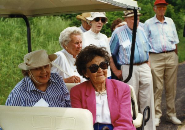 Mary Burke and lifelong friend Jane Matteson sitting in a covered golf cart during a tour near Spring Green, Wisconsin during Mary's 80th birthday celebration. Mary is wearing a safari-type hat and a blue & white stripped blouse. Jane is wearing a pink blazer with a white shirt, sunglasses and a dragonfly necklace. There are seven unidentified people standing in the background.