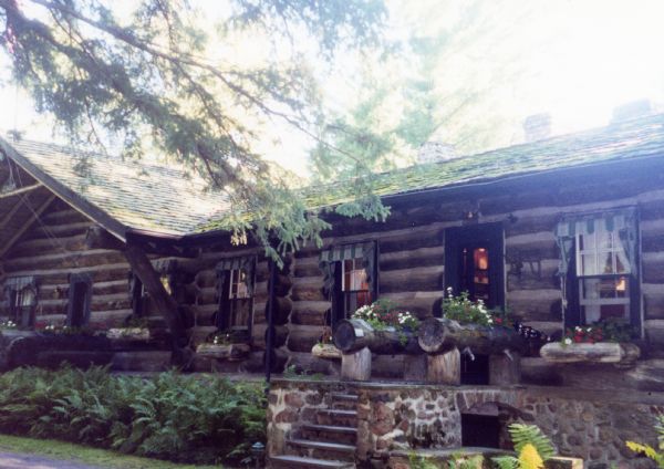 Stone steps leading to the front door of the horizontal chinked log and cedar shake roofed main lodge. Log flower boxes are under the windows and two larger log planters are on each side of the porch. Ferns are along the edge between the house and the lawn.