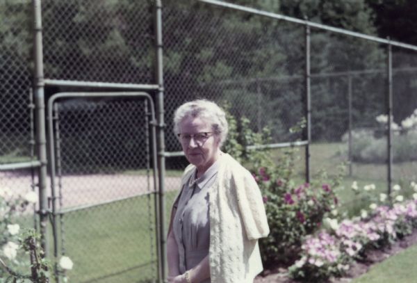Mary Livingston Griggs is standing next to the flowered edged fence of the Forest Lodge tennis court. Mary is wearing a dress with a short-sleeved cardigan over her shoulders.