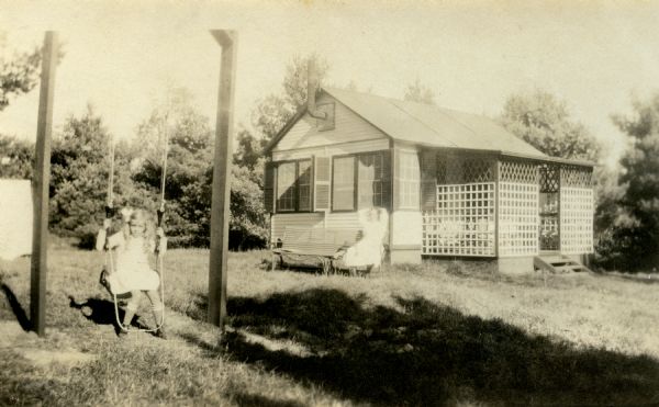 Mary Griggs is sitting on a swing in the lawn near her playhouse. Mary's nurse/nanny is relaxing on a bench against the outside of the playhouse. The playhouse is single story with a lattice porch entrance, windows and a stovepipe protruding near the roof.