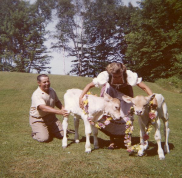 Mary Griggs dressed in a Swedish costume and the dairyman holding two Brown Swiss calves in the pasture near the shore of Lake Namakagon. The calves have flower lei's around their necks.