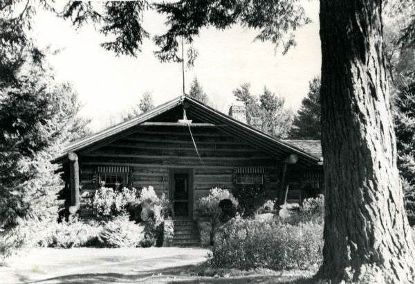 Front doorway to the main lodge which is built of horizontal chinked-logs with stone entrance steps; two stone chimneys on a cedar shake roof. Striped awnings adorn the three front windows. A profusion of flowers are around the entrance and front yard, with the trunk of large pine tree in the immediate foreground.