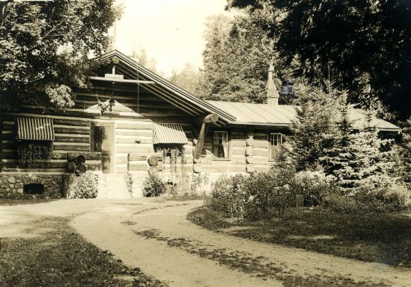 Single-story vertical chinked-log house with a stone foundation and one visible stone chimney. Striped window awnings cover two windows that have window boxes growing ivy. An unpaved driveway curves towards the house next to a flower garden. Many trees are near the garden and house.