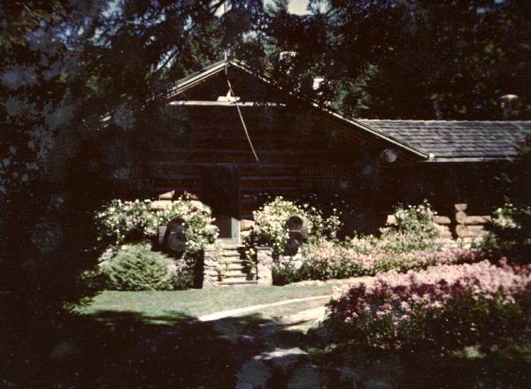 A profusion of flowers grow along the house and front lawn of the main lodge. The lodge is a single-story chinked horizontal log house with a cedar shake roof and stone front porch. A dirt driveway curves between the house and flower garden. Large trees shade a portion of the garden.