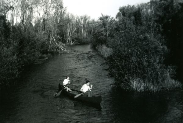 Mary Griggs is sitting in the stern of a canoe paddling the Namakagon River with a male friend sitting in the bow. Mary is wearing shorts and a white blouse, and her male companion is wearing dark slacks and a white shirt.