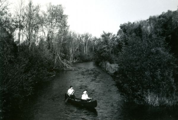 Mary Livingston Griggs is sitting in the bow of a canoe as a young male companion paddles them on the Namakagon River. Mary is wearing a white blouse and hat, her companion is wearing dark trousers and a white shirt. Dense foliage grows along the rivers edge.