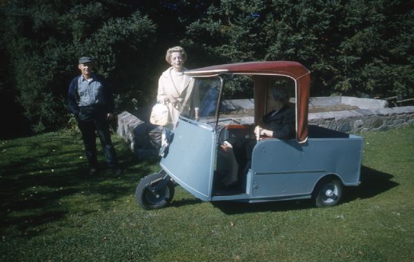 Mary Livingston Griggs, wearing a navy blue dress coat, is sitting in a three-wheel blue cart with a red roof that Mary used to travel the grounds of Forest Lodge. A woman in a pale colored dress and coat, and holding a purse, is standing next to the cart along with a man dressed casually, possibly the caretaker. A stone-walled garden is in the background in front of large spruce trees.