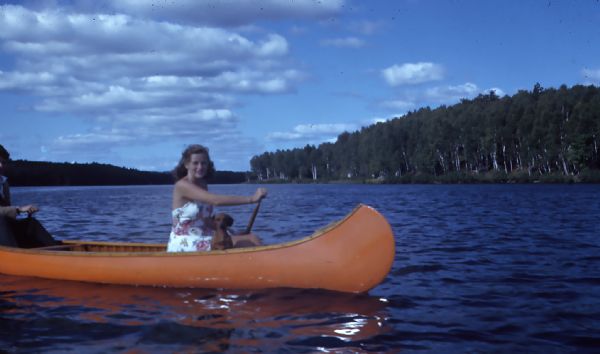 Mary Griggs [Burke] canoeing in a cedar strip canoe on Lake Namakagon with her pet dachshund and a friend. Mary is wearing a floral sundress.