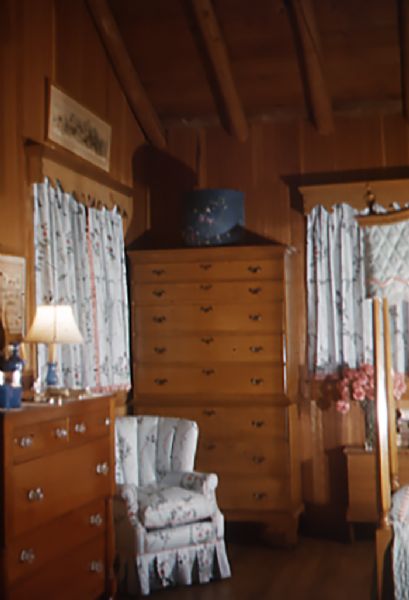 Thin cedar strip paneled room with log ceiling beams. On the left is a pine dresser with a mirror, blue lamp and glass drawer knob. At the back of the room is an eight drawer pine bureau with a blue hat box on top. Other furnishings include floral print chintz fabric curtains, boudoir chair and canopy on four poster bed.