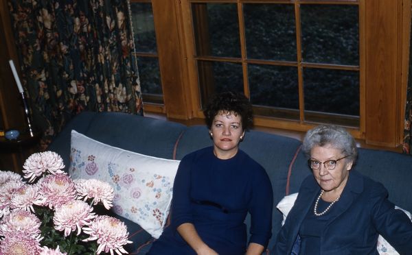 Mary is seated on the right, wearing a navy blue skirt and jacket and a pearl necklace. Her guest is wearing a navy blue dress with three-quarter length sleeves. They are seated in front a large window with floral draperies, on a blue upholstered sofa with embroidered pillows. Pink chrysanthemums are in a vase on a table in front of the sofa. They are in the Great Room of the guest house.